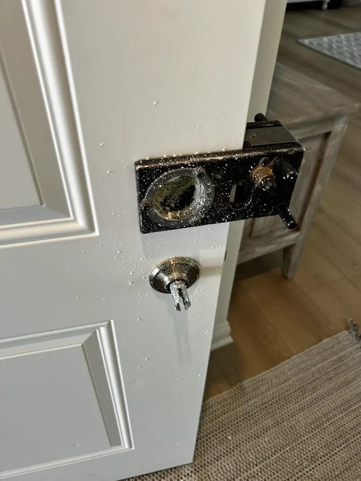 White paneled residential door with an old dark metal plate around a worn opening and a keyed cylinder below it, with debris scattered on the door face