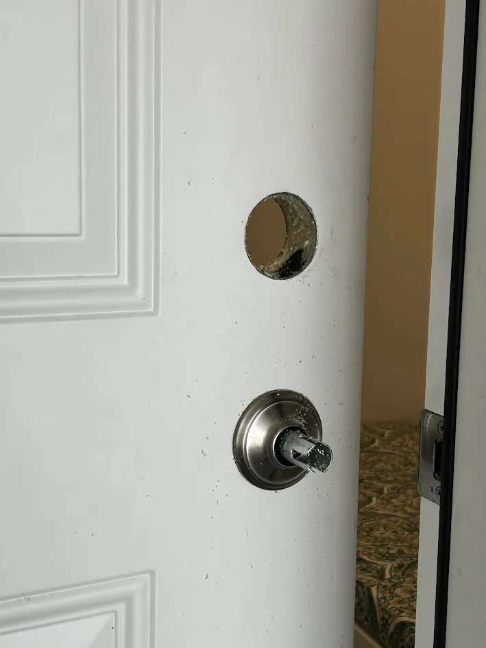 A clean circular cross-bore cut through a white paneled door to take a deadbolt, with the existing keyed lock below it and debris on the door