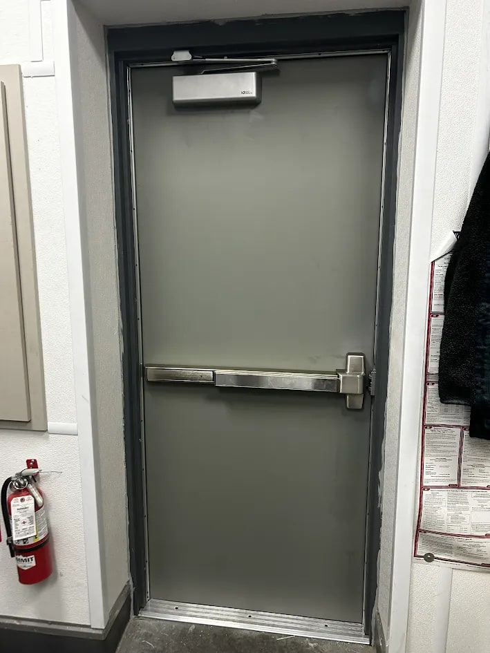 Interior view of the new grey service door showing an overhead door closer and a heavy duty exit bar, with a fire extinguisher on the wall beside it