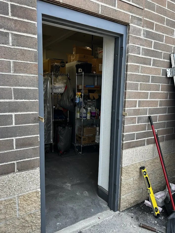 Newly installed dark steel door frame squared and set into the concrete block opening with hinge plates visible and the storeroom beyond