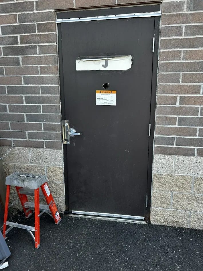 Worn dark hollow metal back service door with a peeling decal and a warning notice taped to it, set in a concrete block wall with a step ladder beside it