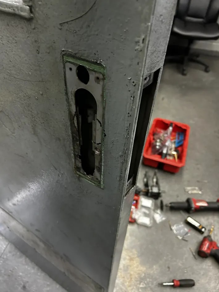 Grey steel door opened at the lock position to expose the mechanism, with tools and a red parts box on the floor