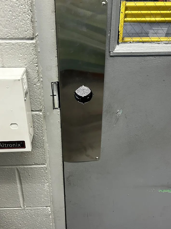 Grey steel commercial door with a long brushed stainless faceplate at the latch and a round black knob through it