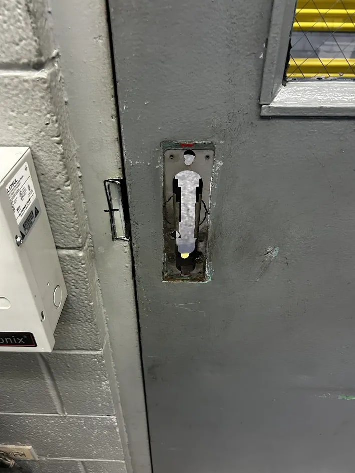 Grey steel commercial door at its latch with a recessed steel plate and vertical slot, a wall-mounted power supply box to the left and a grille in the window above