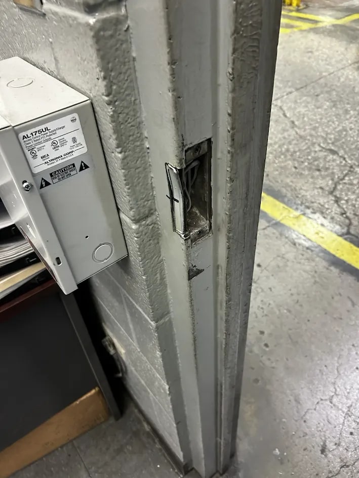 Grey steel door frame with a rectangular mortise cut for an electric strike, wiring inside, beside a wall-mounted power supply enclosure