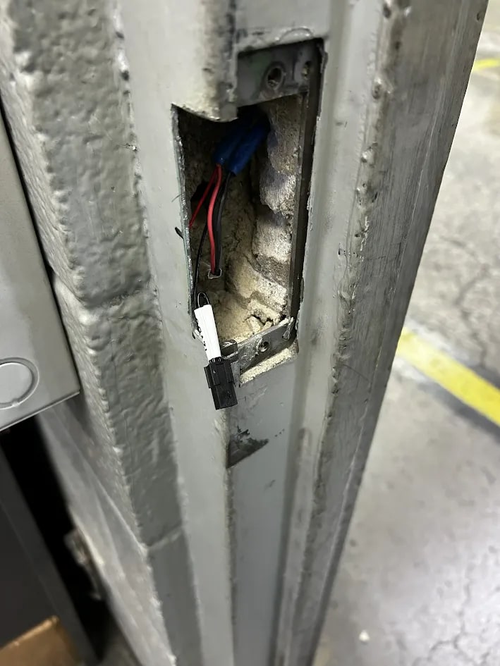 Rectangular cutout in a grey steel door frame with red and black wires and a white connector hanging out