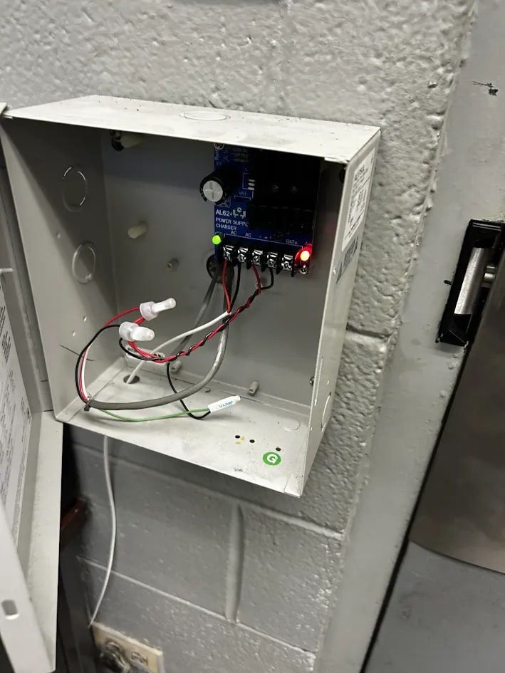 Open power supply enclosure on a block wall with a circuit board, panel wiring and two lit indicator LEDs