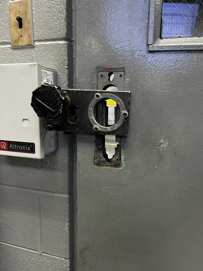 Grey steel door with a black electric strike assembly and round knob at the latch, a galvanised plate beside it and a power supply box on the wall
