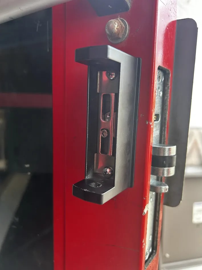 Black rectangular strike assembly mounted on a red steel door frame beside a latch plate on the door edge