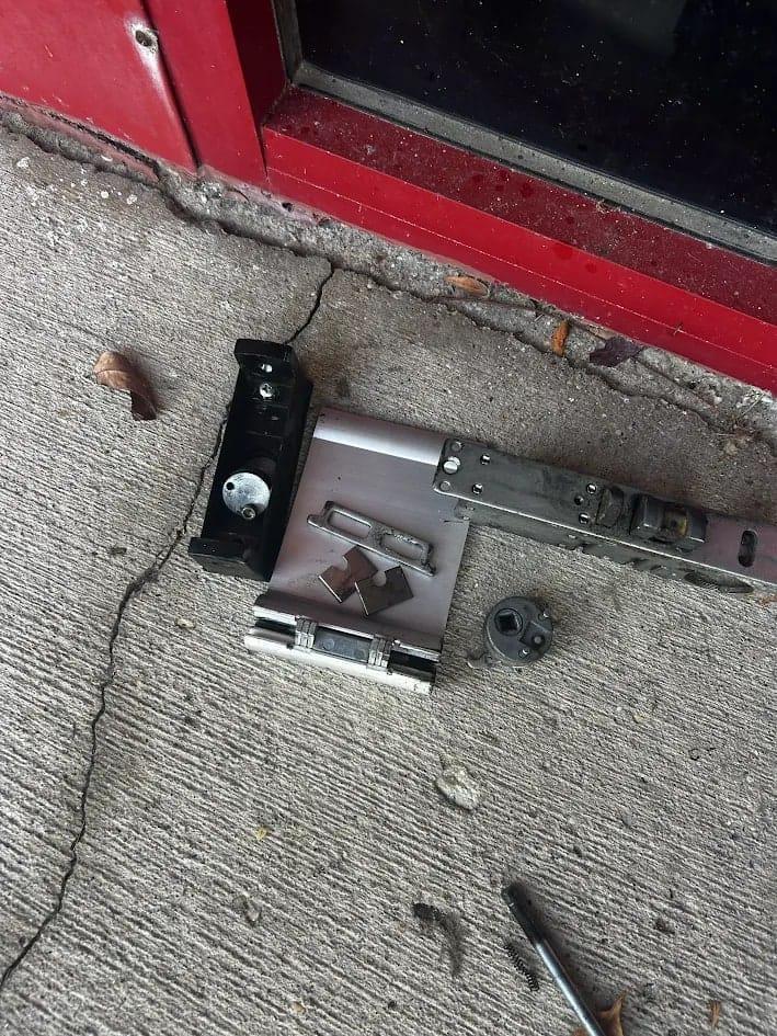 Door hardware laid out on a cracked concrete floor beside a red door, including a lock body, a bracket, a cylinder and fixings