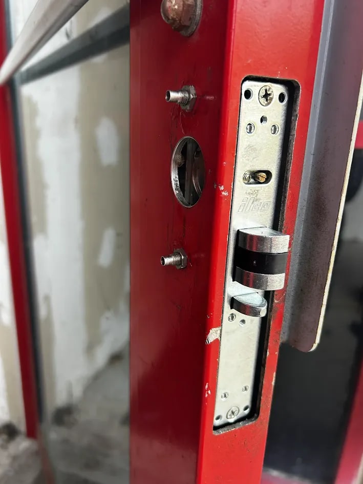 Red-framed glass door with its mortise lock faceplate exposed, threaded mounting studs standing out and an open circular cylinder bore