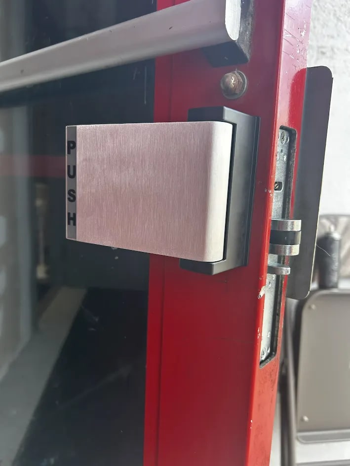 Brushed stainless push paddle marked PUSH mounted on a black backplate on a red-framed glass door